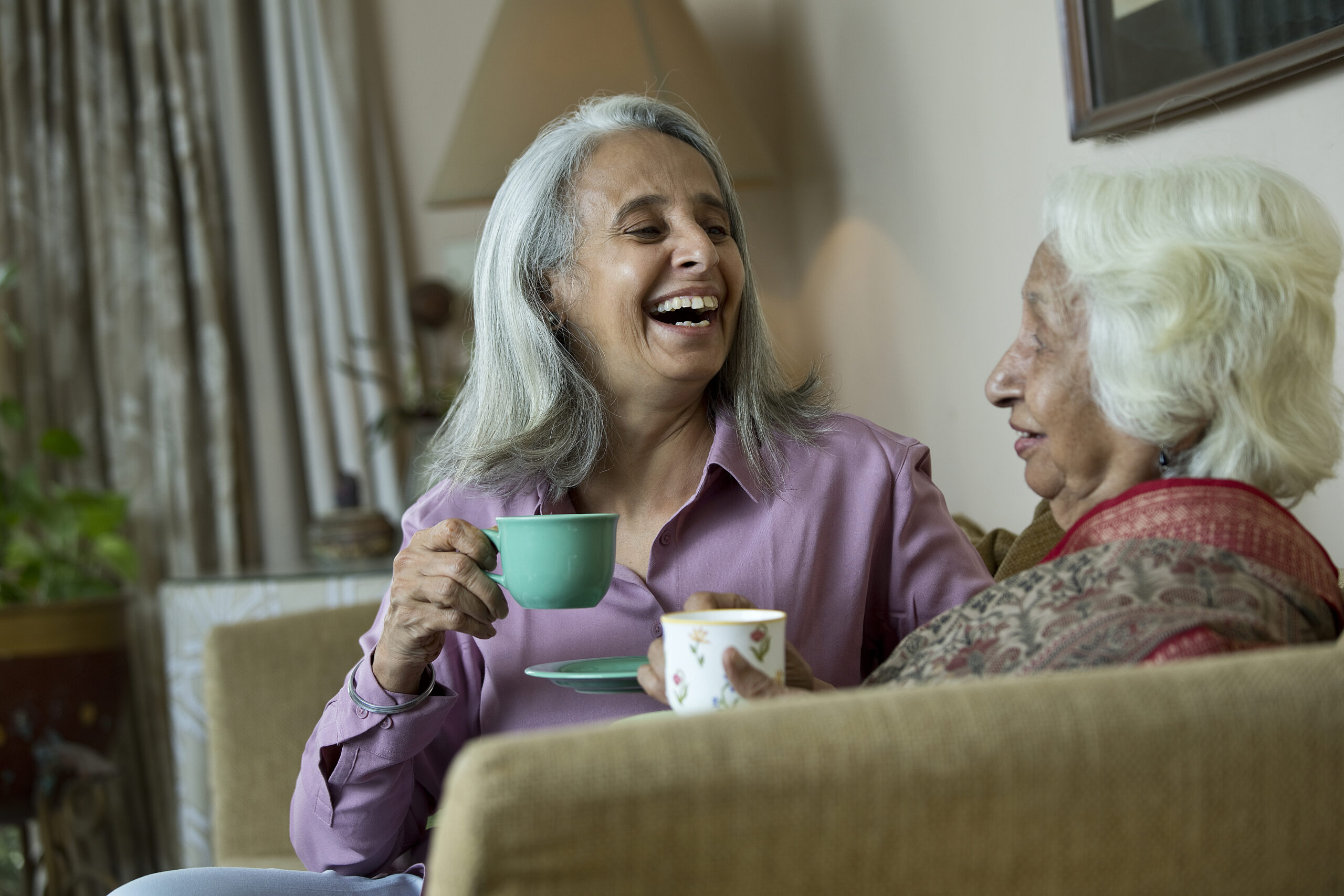 Two old women laughing while drinking coffee at home